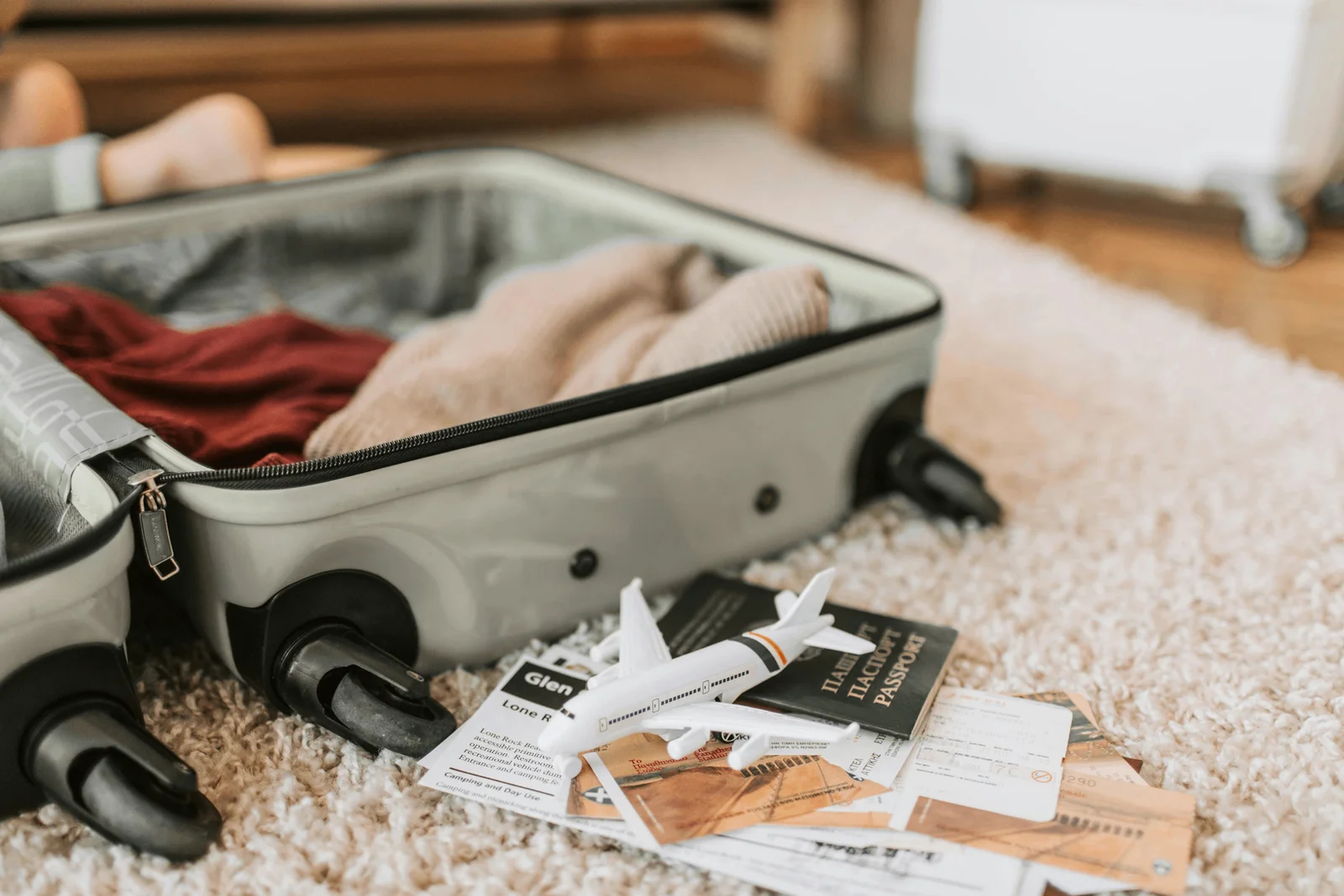 Mala de viagem aberta com roupas organizadas, passaporte, avião de brinquedo, documentos de viagem e acessórios sobre tapete bege, mostrando organização inteligente de bagagem